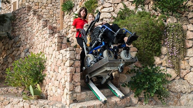 una persona mayor discapacitada en silla de ruedas es transportada por una escalera exterior de piedra con una oruga salvaescaleras eléctrica